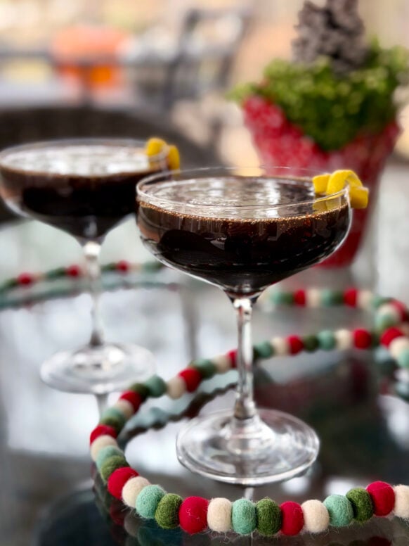 Close-up of ruby-hued Pomegranate French 75 cocktail in coupe glass with delicate foam top, garnished with lemon twist and holiday felt ball garland in foreground