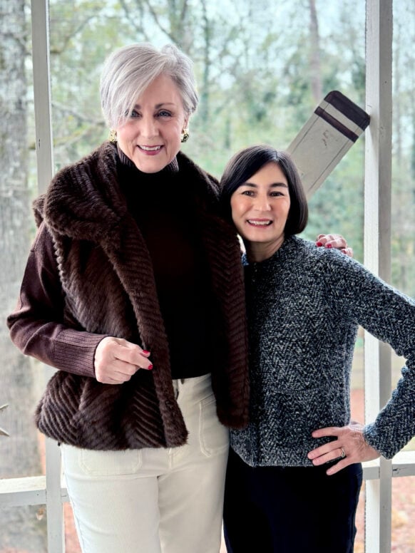 Two women standing together on screened porch, one in luxurious brown faux fur vest over ivory turtleneck with white pants, one in textured charcoal herringbone crop jacket with black barrel-leg pants