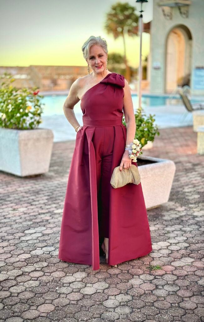 Beth Djalali as Mother of the Groom in a burgundy one-shoulder Alexa Maria jumpsuit with dramatic overskirt, styled with gold mesh clutch and wrist corsage at her son's 2023 wedding