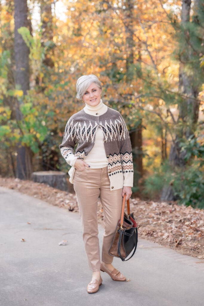 Beth wearing brown and cream fair isle cardigan over cream turtleneck with camel pants, tan loafers, and black leather tote with cognac handles, standing on tree-lined path with fall foliage
