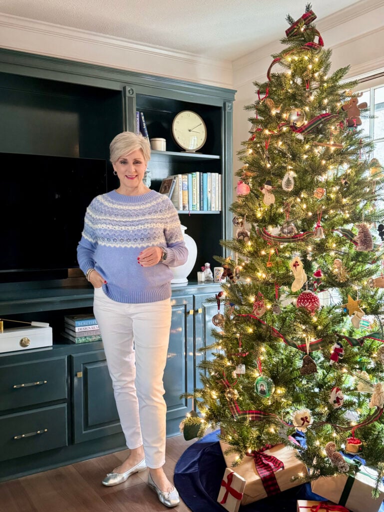 Beth wearing periwinkle blue fair isle sweater with white geometric pattern and matching blue solid sleeves, white ankle pants, silver ballet flats, standing by decorated Christmas tree in living room with teal built-in cabinets