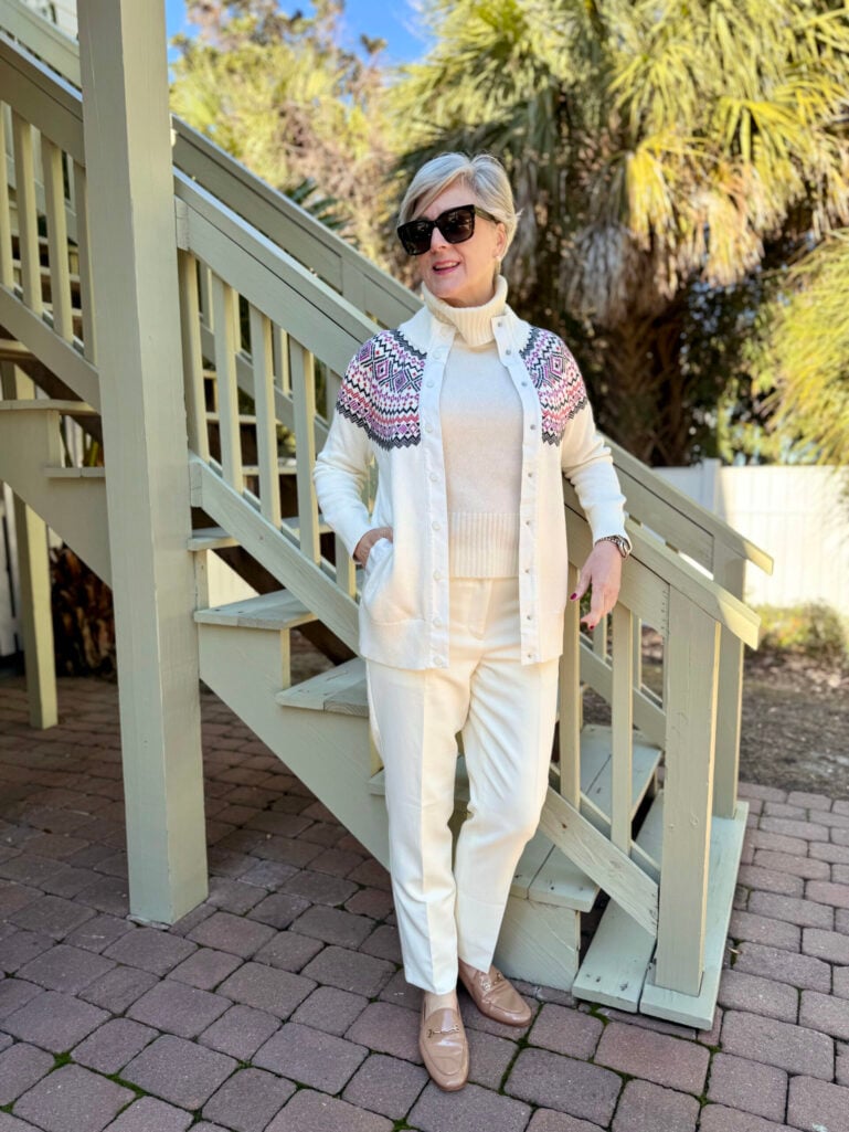 Beth wearing cream fair isle cardigan with pink and black geometric shoulder pattern over cream turtleneck and white pants, tan loafers with gold hardware, oversized sunglasses, standing on paver patio