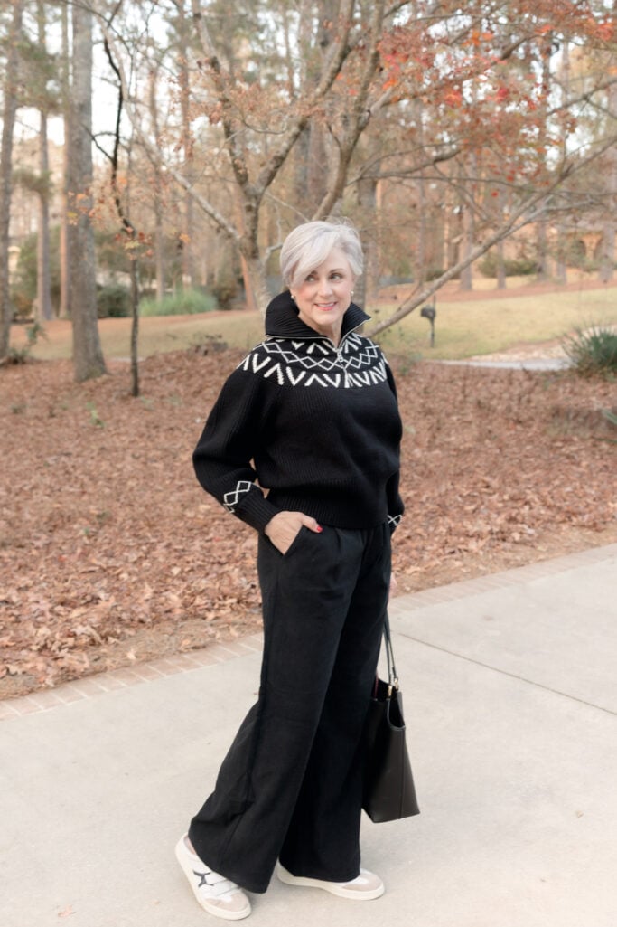 Beth wearing black fair isle quarter-zip sweater with white geometric pattern, black wide-leg pants, white sneakers, and black shoulder bag, standing on sidewalk with fall trees in background