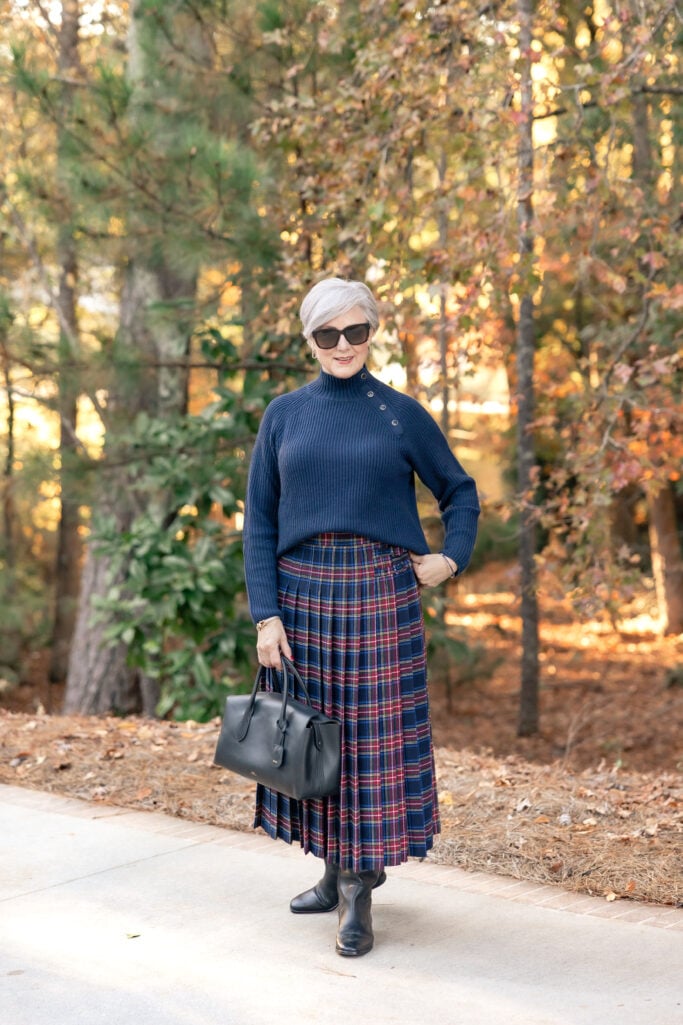 Beth in a navy and red tartan pleated kilt skirt with a navy button-neck sweater, black ankle boots, black leather satchel, and oversized sunglasses—classic tartan kilt outfit over 50