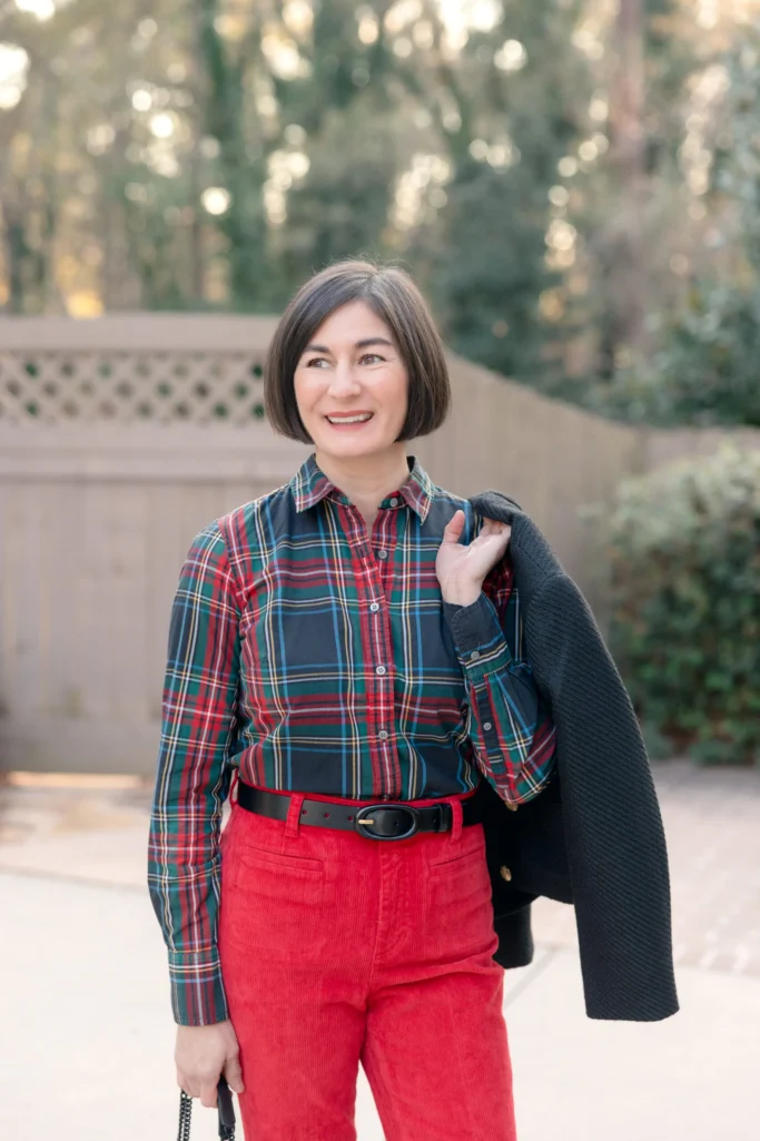 Woman wearing a green and red Stewart tartan button-down shirt tucked into red corduroy pants with a black leather belt and navy jacket draped over shoulder—casual holiday plaid outfit