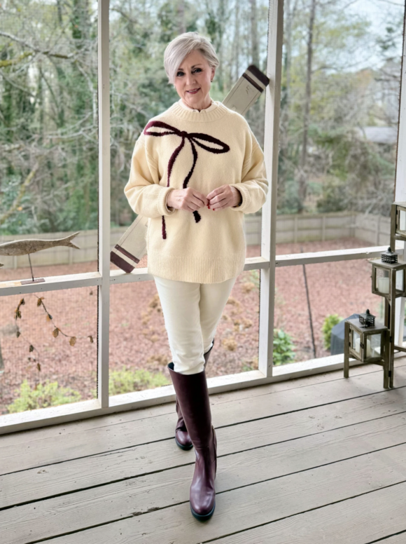 Woman with short silver hair wearing a cream sweater with a bow detail, white pants, and tall burgundy boots, standing on a screened porch with a wooded backyard view, styled for a polished yet cozy seasonal outfit.