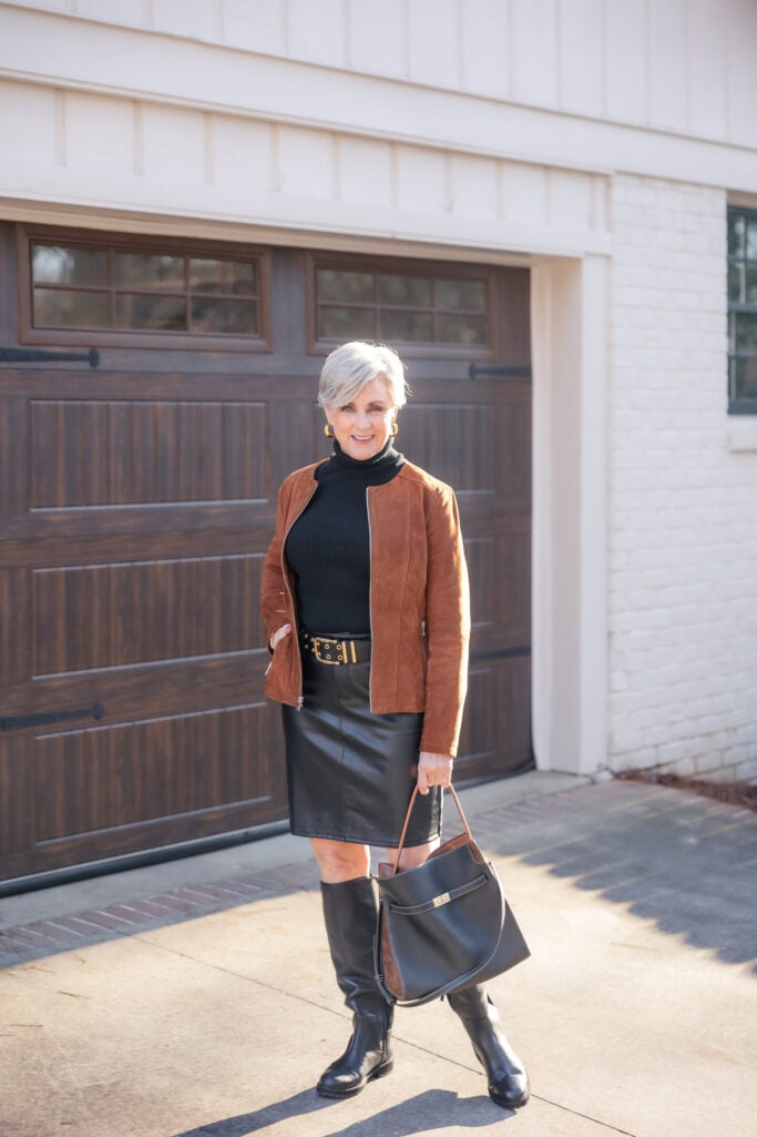 Woman over 50 wearing cognac suede moto jacket with black turtleneck, leather skirt, and knee-high boots