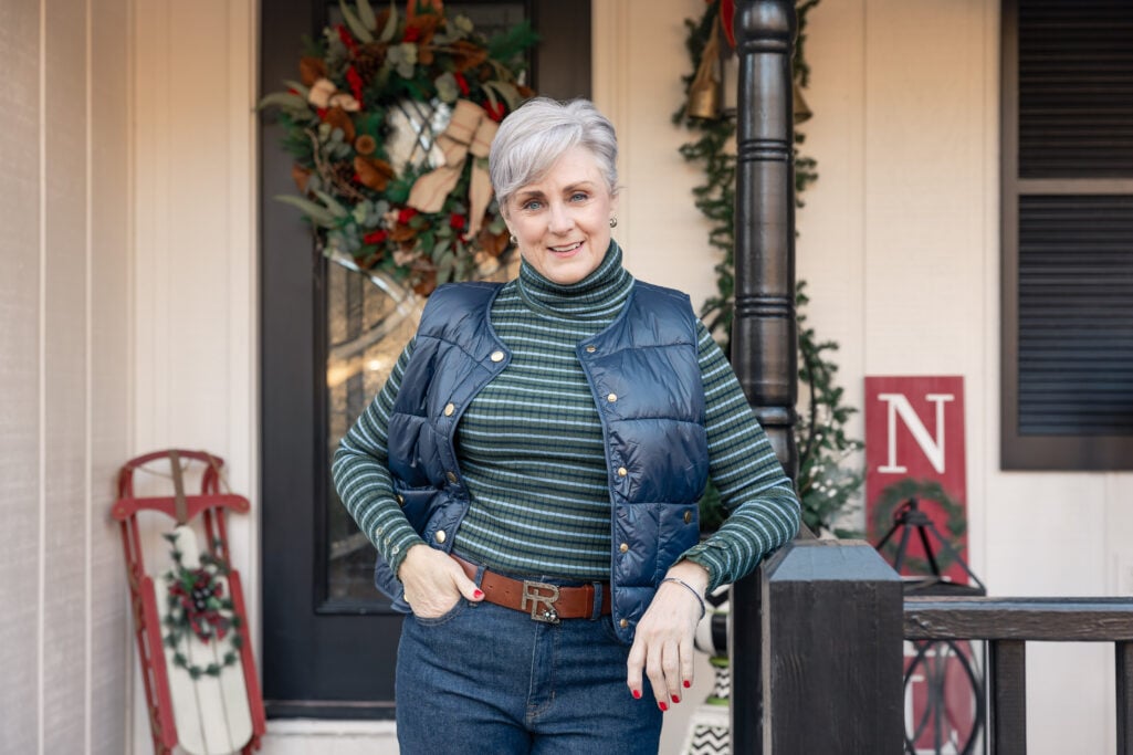 Beth wearing a green striped turtleneck with a navy puffer vest and jeans with a cognac leather belt, standing on a festively decorated porch with holiday wreaths and a vintage red sled—casual winter outfit inspiration