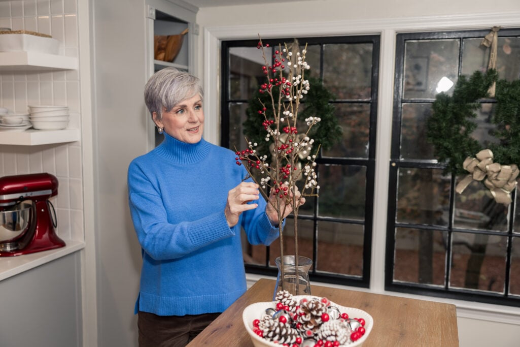 Beth in a bright blue cashmere turtleneck sweater arranging red and white berry branches in her kitchen, with holiday pinecone decor and a wreath on the window—cozy cashmere sweater gift idea for the holidays
