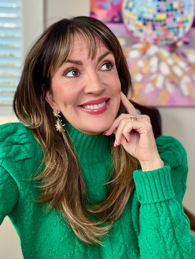 Woman smiling with finished makeup look showing rosy cheeks, glossy berry lips, and shimmery eyes, wearing emerald green cable knit sweater and gold celestial star drop earrings, hand posed near chin with candy cane nail art