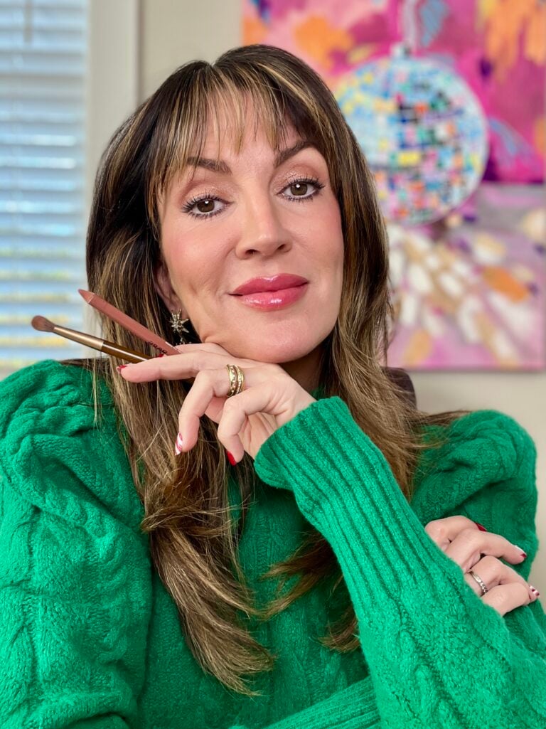 Woman in green turtleneck sweater holding nude lip liner and makeup brush near chin, wearing shimmery eyeshadow and glossy lips, gold star earrings visible, colorful disco ball artwork in background