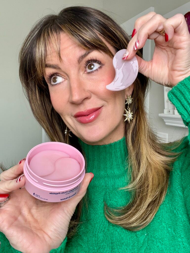 Woman holding pink Terez and Honor collagen eye patch near under-eye area while displaying pink jar of K-beauty eye patches, wearing green sweater and gold star drop earrings with candy cane manicure