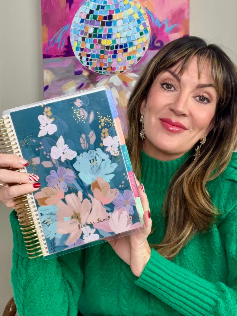 Woman smiling holding personalized Erin Condren Life Planner with teal floral cover featuring her name "Jennifer Duvall," gold coil binding visible, colorful disco ball painting in background