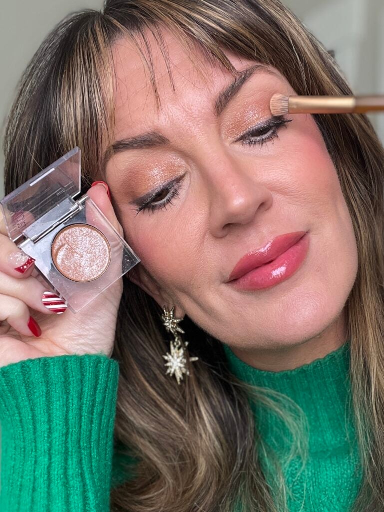 Woman applying Urban Decay Moondust eyeshadow in Space Cowboy with brush while holding open compact, showing warm champagne sparkle shadow on lids, wearing gold star earrings and green sweater
