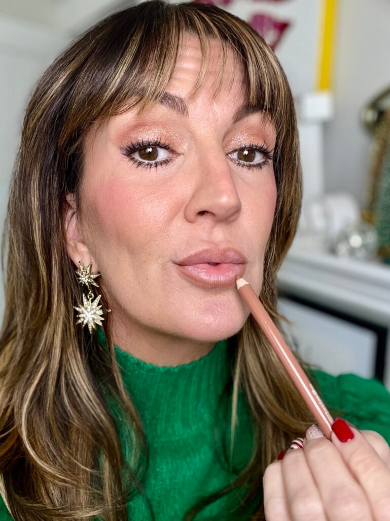 Woman applying nude e.l.f. Creamy Glide lip liner in Baddest Beige to lips, showing shimmery eye makeup and gold starburst earrings, green turtleneck sweater visible