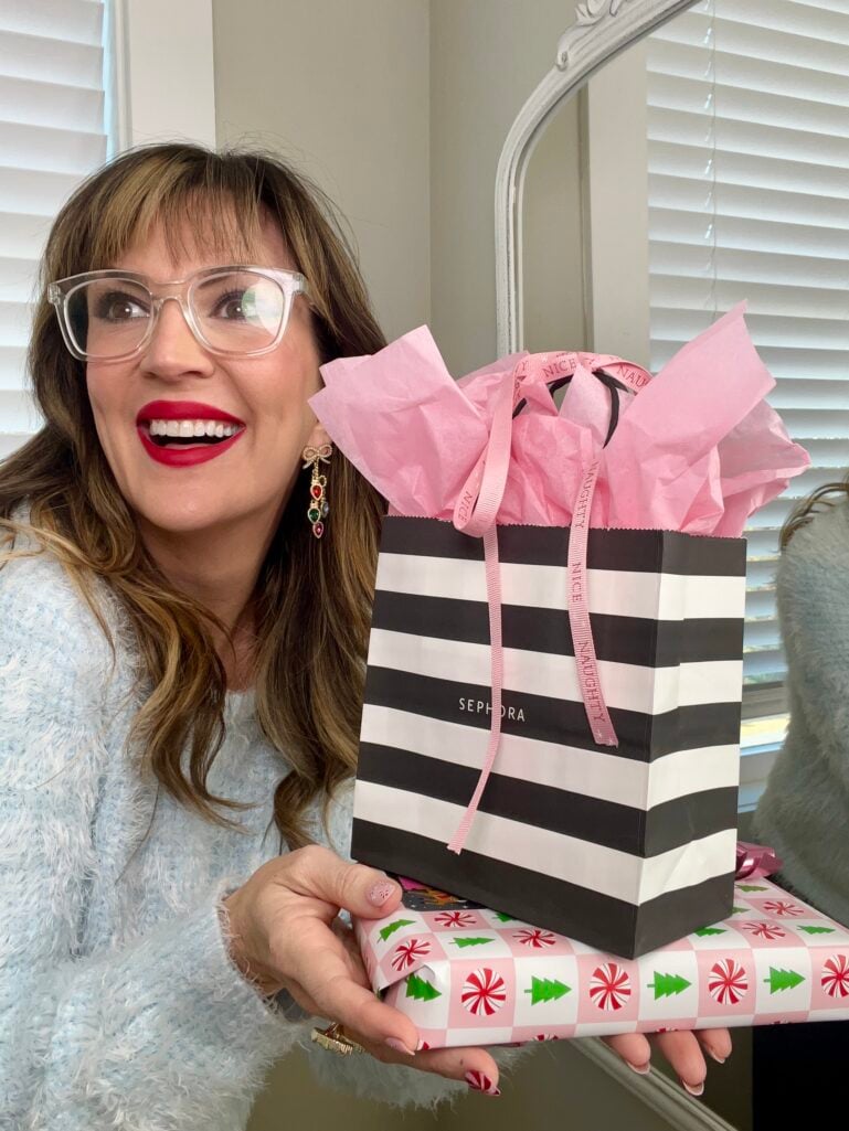 Woman wearing ice blue fuzzy sweater and clear glasses holding Sephora striped gift bag and wrapped holiday present with candy cane nails