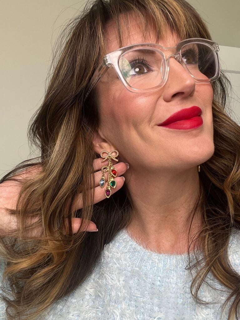 Close-up of woman wearing clear Peepers readers and colorful gemstone chandelier earrings with red liquid lipstick