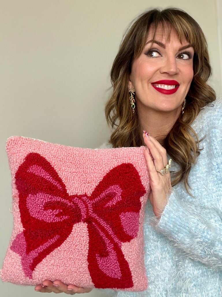 Woman holding pink and red needlepoint pillow with decorative bow design wearing ice blue fuzzy sweater and colorful drop earrings