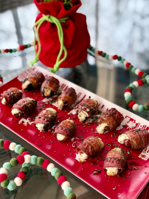 Bacon-wrapped dates with goat cheese and balsamic glaze arranged on a red holiday platter with felt ball garland