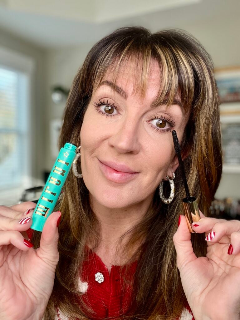 Woman holding Milani highly rated lash extensions tubing mascara and lash curler with one eye showing mascara application