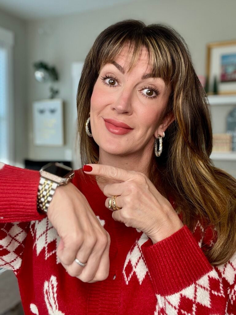 Woman pointing to two-tone gold and silver watch on wrist wearing red holiday cardigan showing natural polished makeup