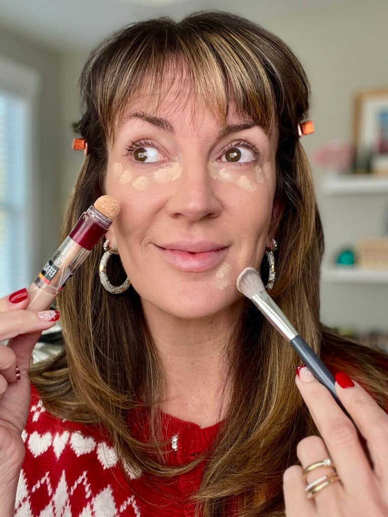 Woman with concealer dots under eyes around nose and on eyelids holding Maybelline Instant Age Rewind concealer and makeup brush