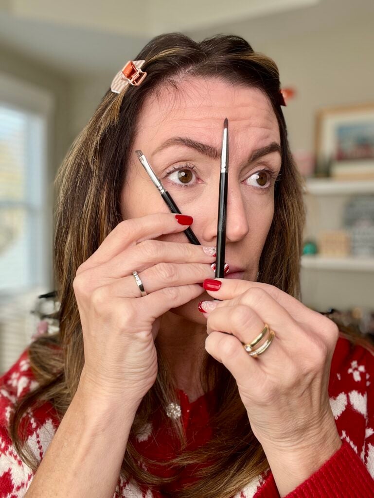 Woman demonstrating eyebrow measuring technique holding pencil and brush vertically to show where brows should start and end