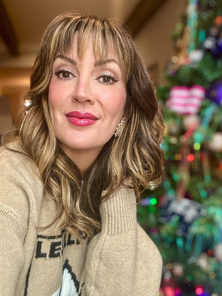 Woman with highlighted brown hair wearing beige Les Alpes sweater and statement crystal earrings, full holiday makeup with berry lips in front of decorated Christmas tree