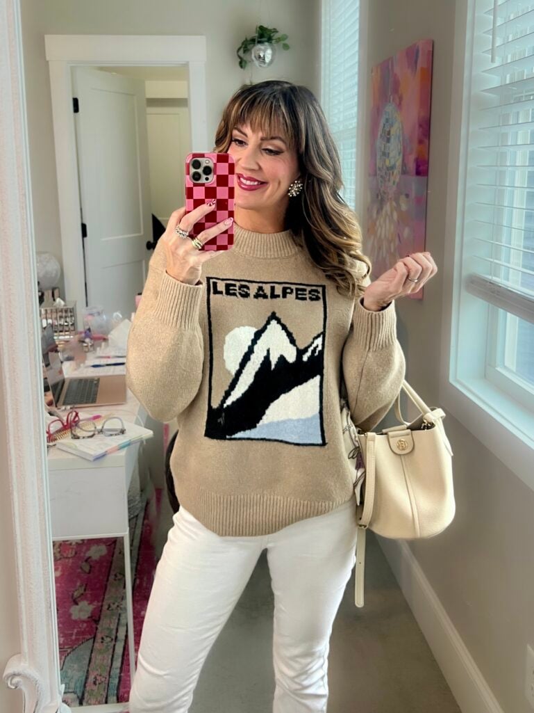 Full-length mirror selfie showing woman in beige Les Alpes mountain sweater, white jeans, bucket bag, complete holiday makeup with candy cane nails