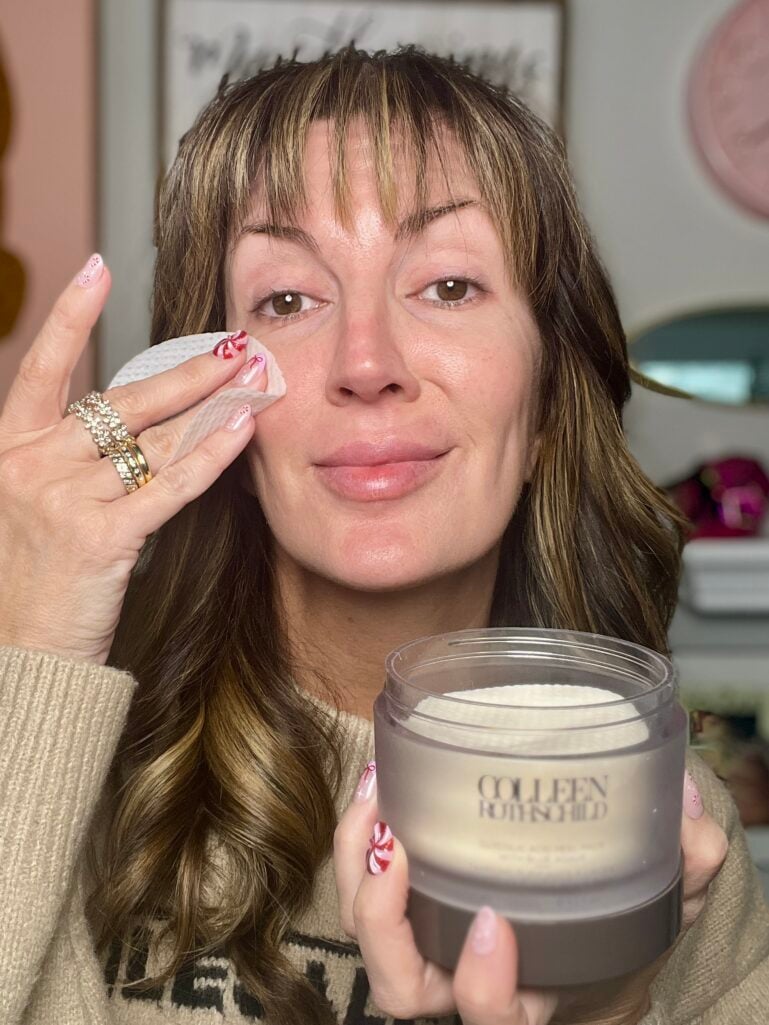 Woman with candy cane nail art applying Colleen Rothschild glycolic acid peel pad to bare face, wearing beige graphic sweater