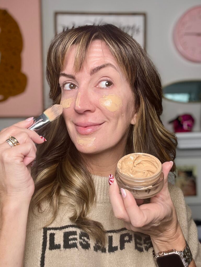 Woman applying Jones Road What The Foundation with makeup brush, showing product swatches on cheeks and nose