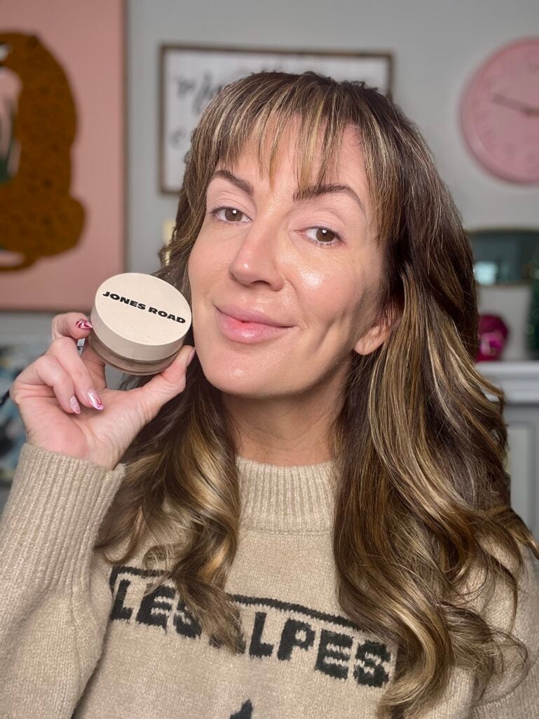 Woman holding Jones Road foundation jar with radiant glowing makeup applied to one side of face