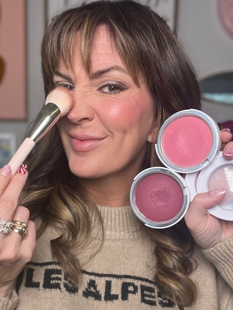 Woman applying cream blush with brush while holding two Makeup by Mario Soft Pop blushes in pink and berry shades