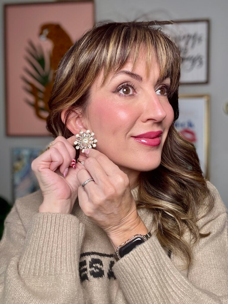 Close-up of woman adjusting large crystal floral statement earring, showing rosy cream blush and glossy berry lip