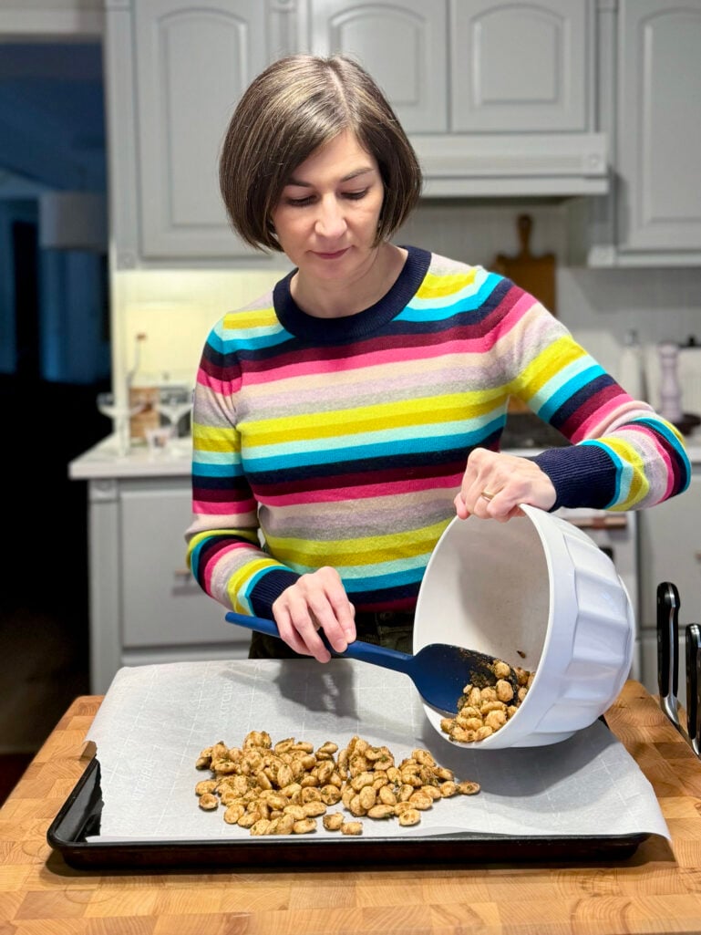 Kelly in a colorful striped sweater spooning spiced Marcona almonds from a white mixing bowl onto a parchment-lined baking sheet