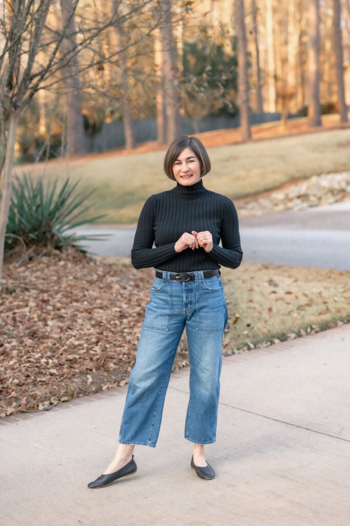 Kelly smiling in petite barrel leg jeans outfit showing cropped ankle length and patch pocket detail - black turtleneck tucked in with belt