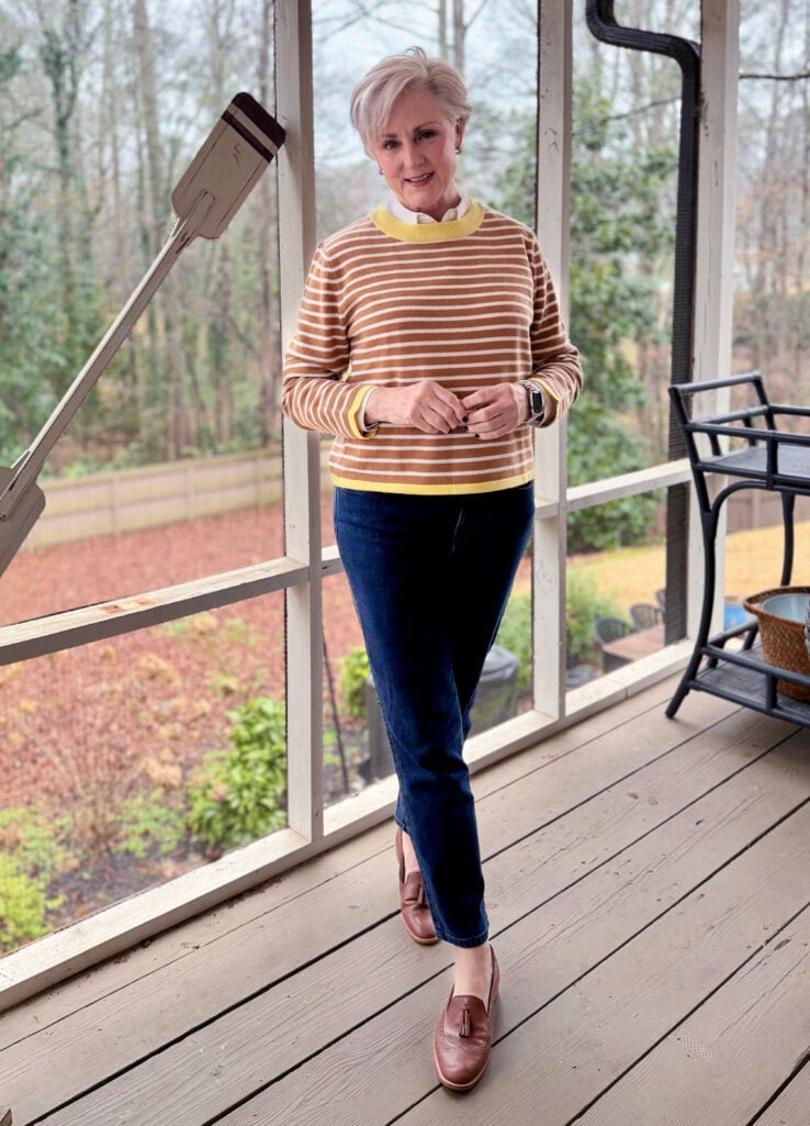 Beth wearing a camel and white striped cashmere sweater with yellow trim over a white button-up shirt, dark jeans, and blush tassel loafers on the screened porch