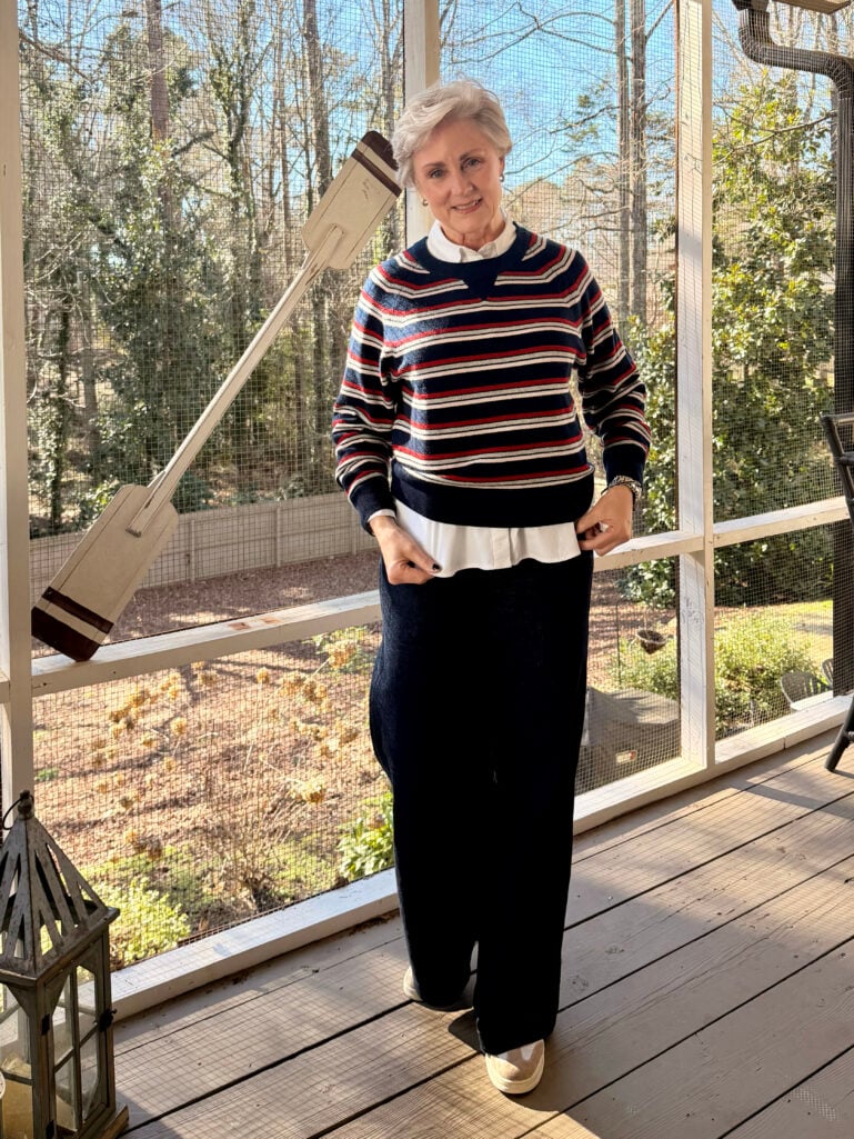 Beth wearing a navy, red, and cream striped cashmere crewneck sweater layered over a white no-iron button-up shirt, wide leg indigo jeans, and cream mule sneakers on the screened porch