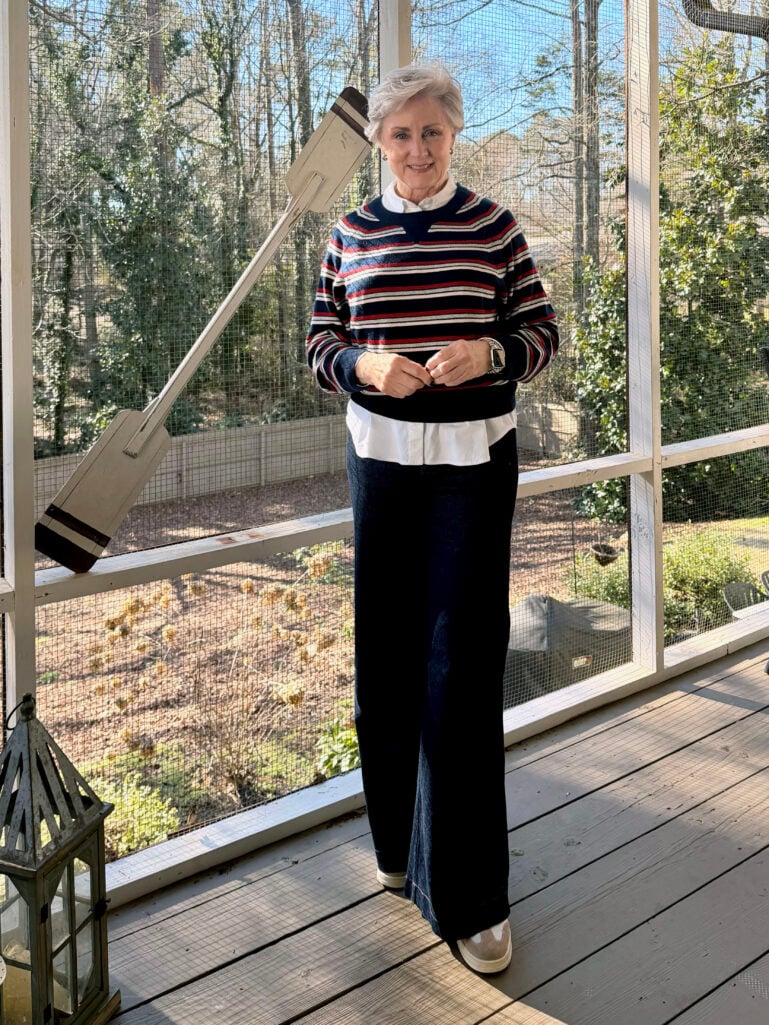 Full-length view of Beth in her striped cashmere sweater and Spanx wide leg jeans, hands clasped, winter trees visible through the porch screens