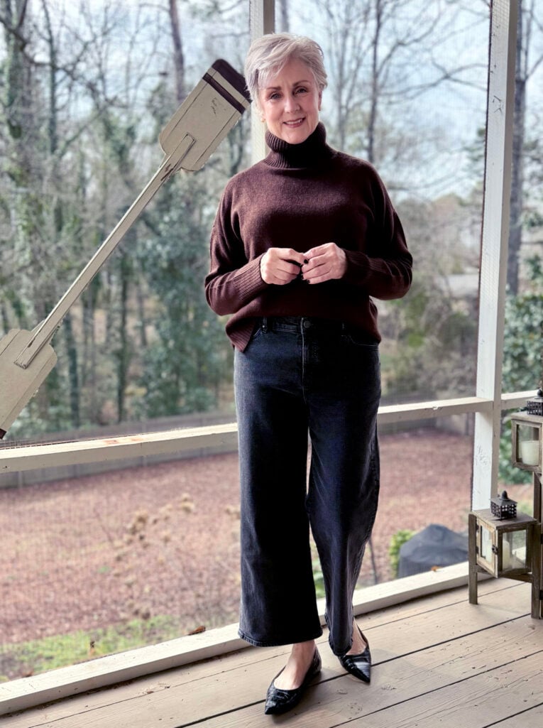 Beth wearing a chocolate brown turtleneck sweater, wide leg black denim, and patent croc-embossed flats on the screened porch