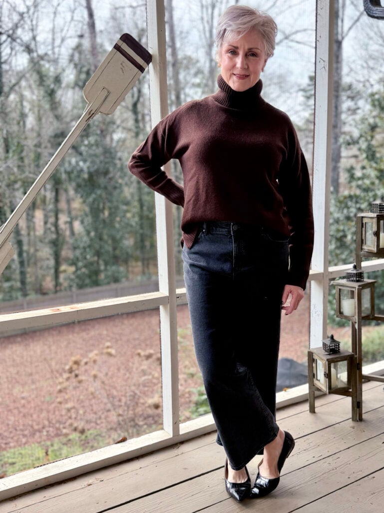 Full-length view of Beth in her brown turtleneck and black wide leg jeans, hand on hip, winter trees visible through the porch screens