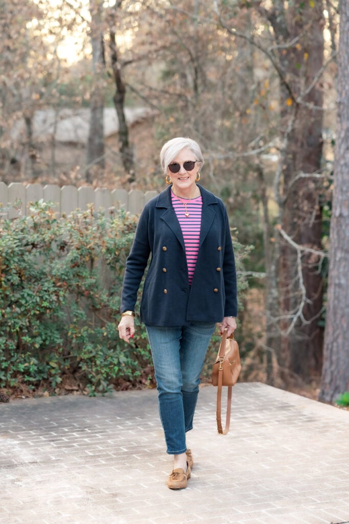 Woman wearing a navy double-breasted jacket with gold buttons over a pink striped top, straight-leg jeans, and tan loafers, carrying a tan crossbody bag outdoors.