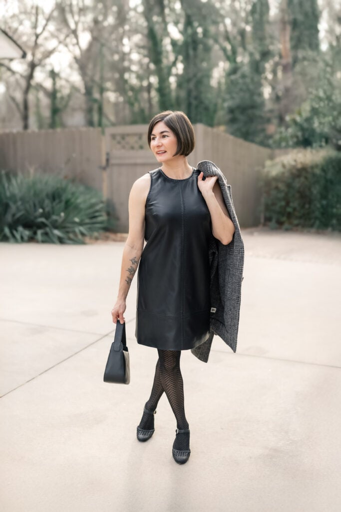 Kelly walking in black faux leather mini dress with grey plaid long jacket over shoulder, herringbone tights and black woven heels - full length petite outfit
