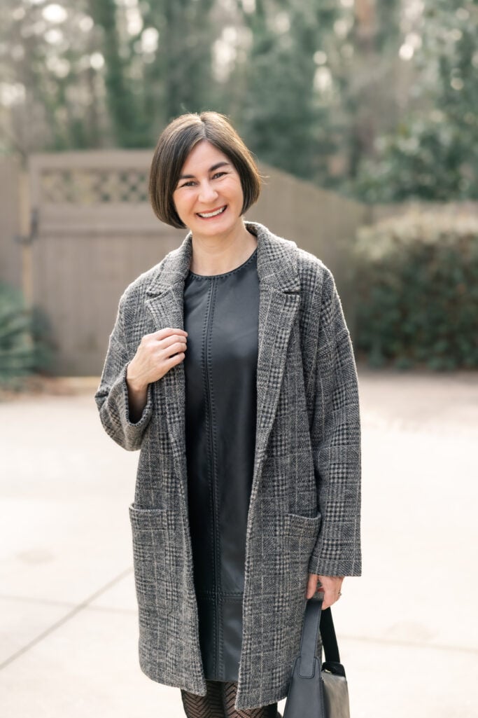 Kelly smiling in black faux leather dress and grey plaid long knit jacket - upper body shot showing texture mixing with leather and knit fabrics