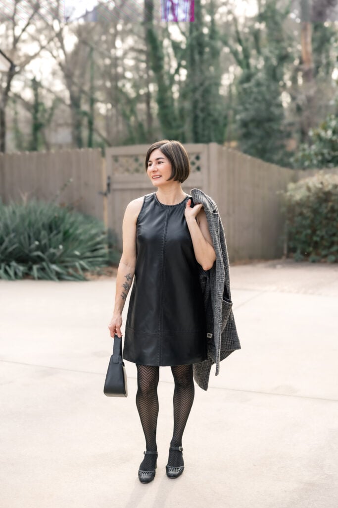 Kelly holding grey plaid coatigan over shoulder showing black faux leather sleeveless mini dress with herringbone tights and woven heels - petite dress outfit without jacket