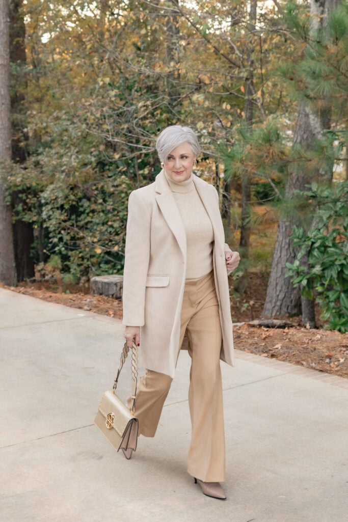 Beth wearing monochromatic camel outfit with wide-leg trousers, cashmere turtleneck, open-front coat draped over shoulders, taupe pointed-toe heels, and Tory Burch chain-strap handbag with gold hardware
