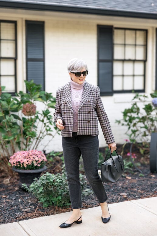 Woman with short gray hair wearing a cropped plaid jacket over a blush pink cable-knit turtleneck, black straight-leg jeans, and black slingback flats, carrying a structured black handbag while walking outside in front of a white house with black shutters.