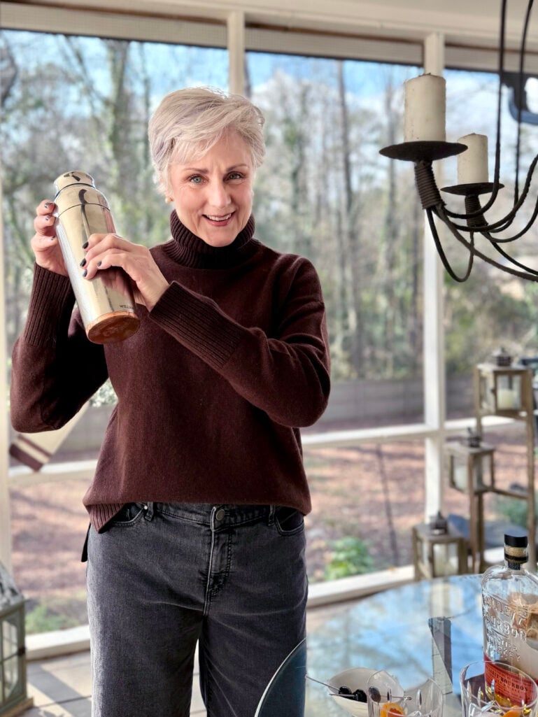 Beth smiling while shaking a cocktail in a silver shaker, wearing her chocolate brown turtleneck on the screened porch