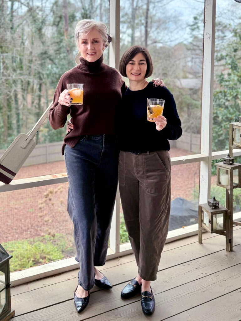Beth and Kelly raising blood orange whiskey sours on the screened porch, showing their full outfits with winter trees visible through the windows