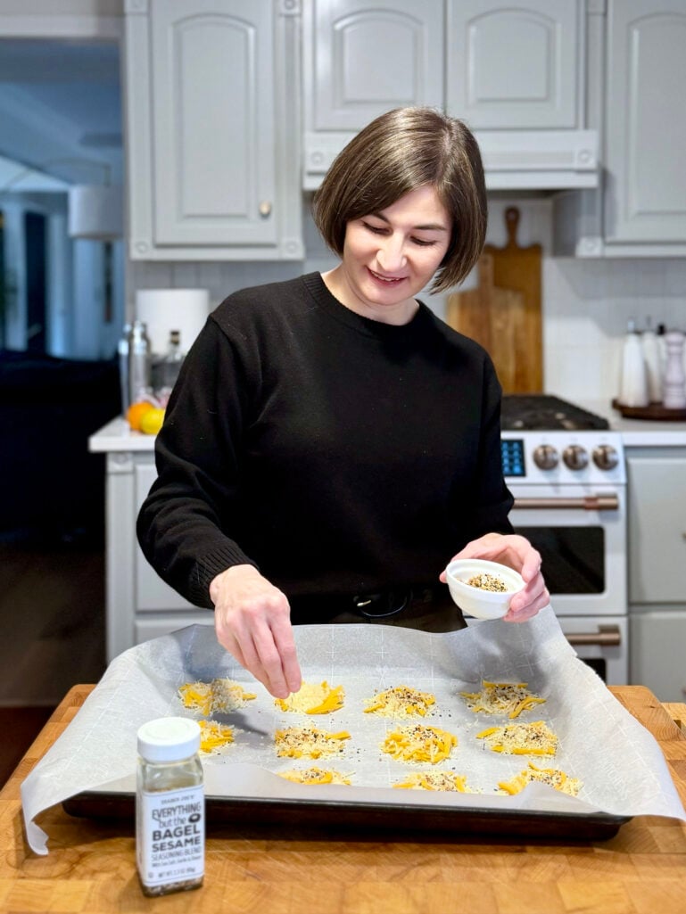 Kelly sprinkling everything bagel seasoning over shredded cheese mounds on a parchment-lined baking sheet, Trader Joe's Everything But the Bagel seasoning jar visible